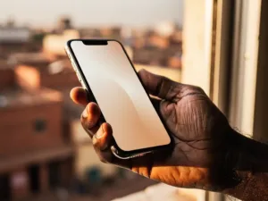 Weathered hand holding a modern smartphone with an African cityscape softly blurred in the background, bathed in warm golden window light.