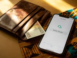 Worn leather wallet with euro banknote beside a smartphone showing money transfer confirmation, resting on kente-patterned fabric in warm amber light.