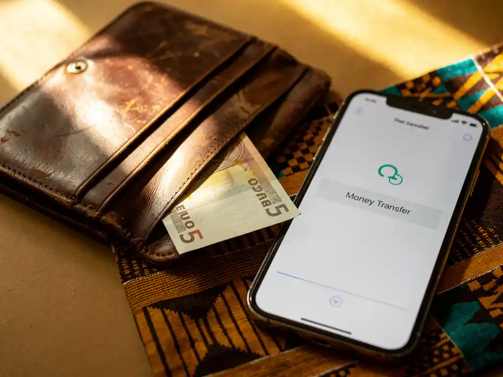 Worn leather wallet with euro banknote beside a smartphone showing money transfer confirmation, resting on kente-patterned fabric in warm amber light.