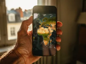Weathered hand holding a smartphone displaying a glowing world map with connection arcs linking Europe and Africa, warm afternoon light.