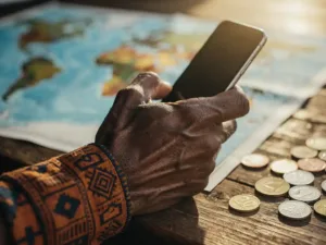 Weathered hand holding a smartphone over a wooden desk with a blurred world map and scattered multi-currency coins in warm golden light.