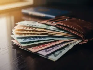 Fanned international currency notes on a leather travel wallet beside a passport, warm golden light on a wooden surface.
