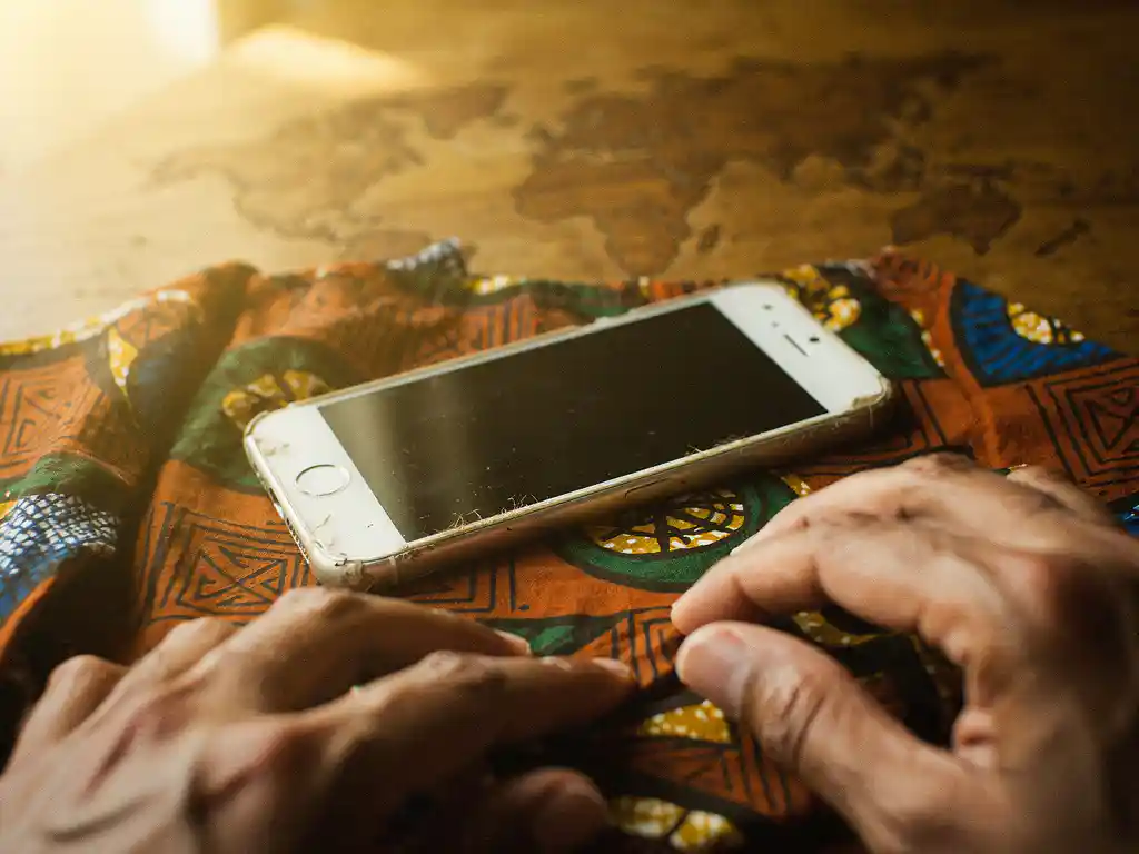 Worn iPhone on colorful African wax-print fabric over a wooden world map, warm hands adjusting settings in soft golden light.