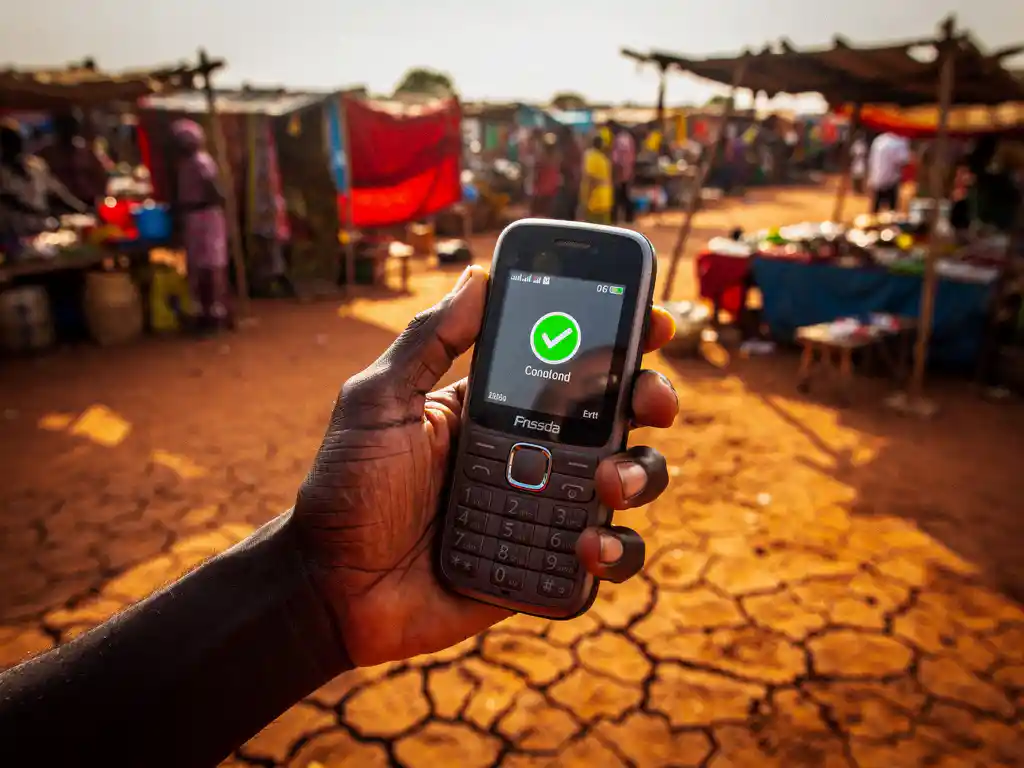 Weathered hand holding a basic mobile phone showing a transaction confirmation screen at a South Sudanese open-air market in warm afternoon light.