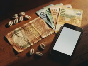 Weathered Nigerian naira and euro bills on a wooden surface with cowrie shells and a glowing smartphone nearby.