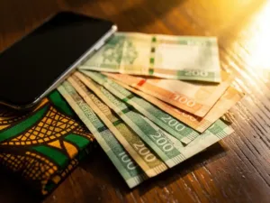 Nigerian naira banknotes fanned out beside a smartphone on a wooden surface with an Ankara fabric swatch, bathed in warm golden light.