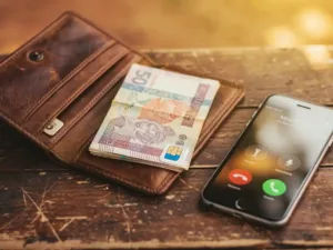 Weathered leather wallet open on wooden table with Sudanese pound banknotes, prepaid SIM card, and smartphone showing an active international call.