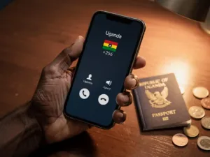 Weathered hand holding a smartphone with an incoming call showing a Ugandan +256 indicator, beside a passport and coins on a wooden desk.