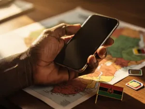 Weathered hand holding a smartphone over a blurred Uganda map on a wooden desk, with scattered SIM cards and a small Ugandan flag pin.