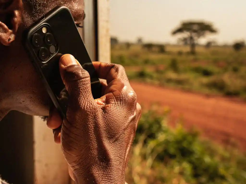 Weathered hand holding a smartphone to the ear, with a sunlit Ugandan landscape softly blurred in the background.