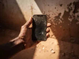 Weathered hand holding a cracked, unresponsive smartphone on a dusty Eritrean street, afternoon sunlight cutting across a peeling concrete wall.