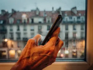 Weathered hand holding a smartphone to the ear, rain-streaked window framing a blurred European cityscape in warm amber and slate blue tones.