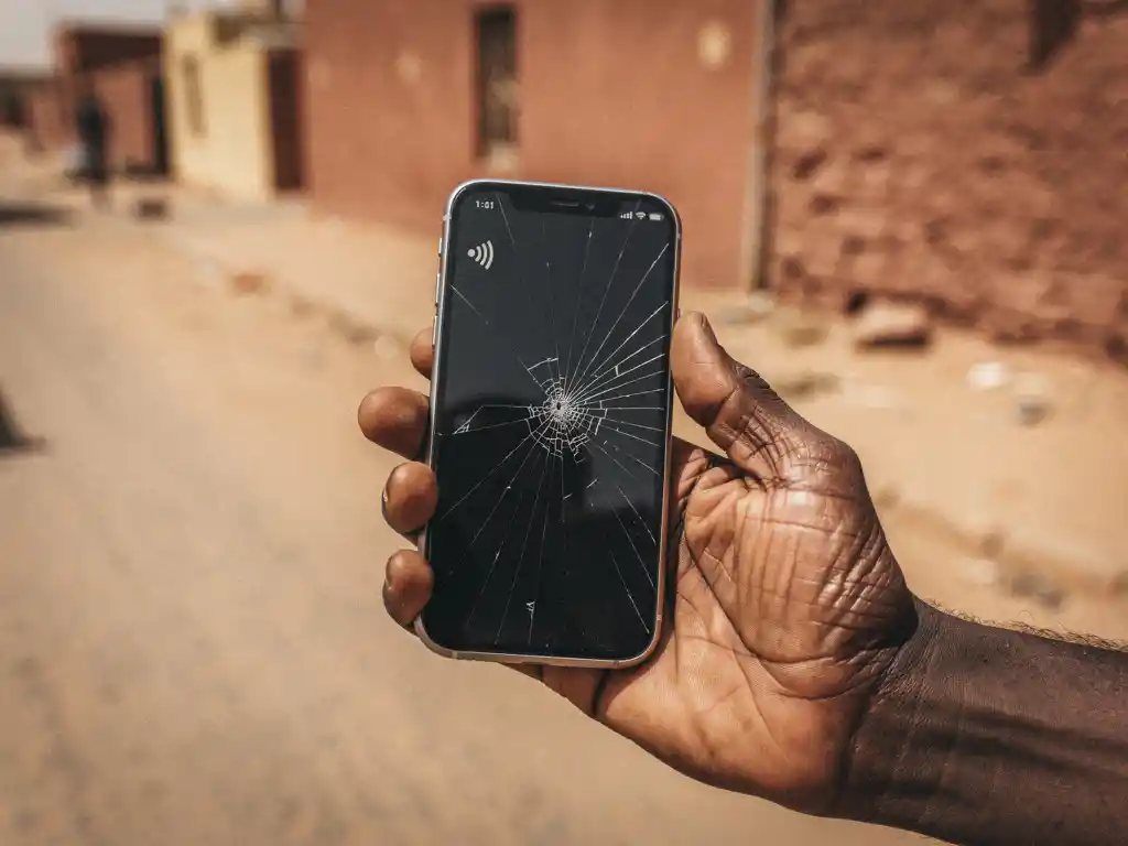 Weathered hand holding a cracked smartphone on a sun-baked Sudanese street with dusty terracotta walls and golden midday light.