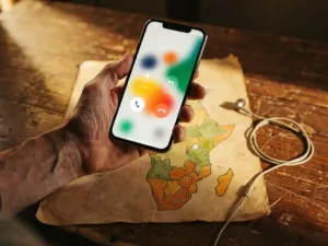 Weathered hand holding a smartphone on an active call over a hand-drawn map of Africa, with a coiled earphone on a worn wooden surface.