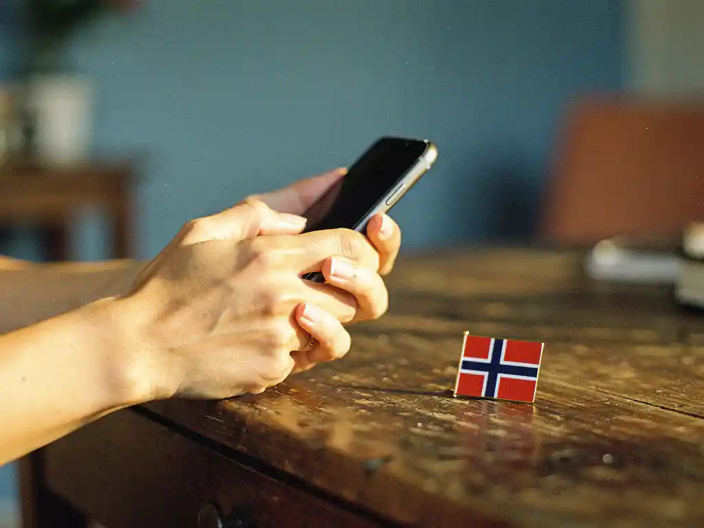 Hands holding a smartphone mid-call on a wooden desk beside a Norwegian flag pin, warm amber tones blending with Nordic blue light.