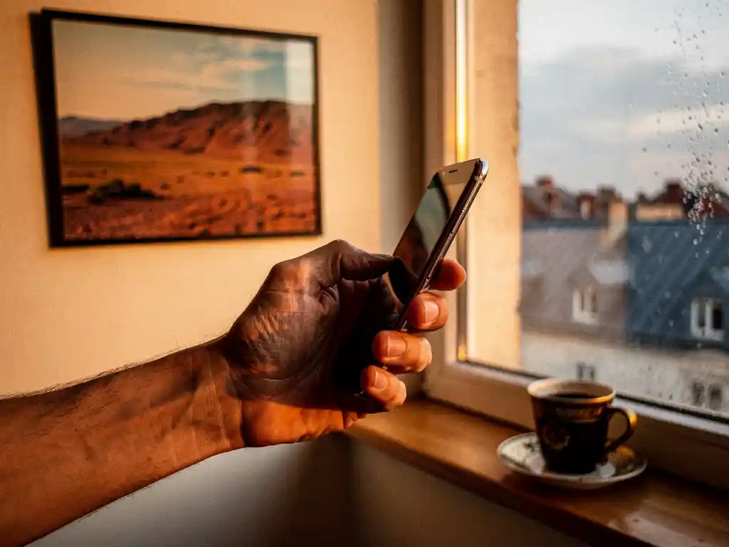 Weathered hand holding a smartphone in a warmly lit apartment, Eritrean landscape photo and Ethiopian coffee cup softly blurred on a rain-streaked windowsill.