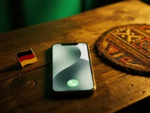 Smartphone mid-call on a weathered wooden desk beside a German flag pin and Tanzanian woven textile coaster in warm golden light.