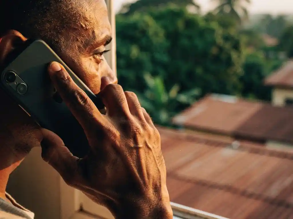 Weathered hand holding a smartphone to an ear near a sunlit window overlooking green Ghanaian rooftops in warm amber light.