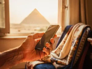 Weathered hand holding a smartphone near a sunlit window with a faint pyramid silhouette, Arabic textile draped alongside in warm terracotta tones.