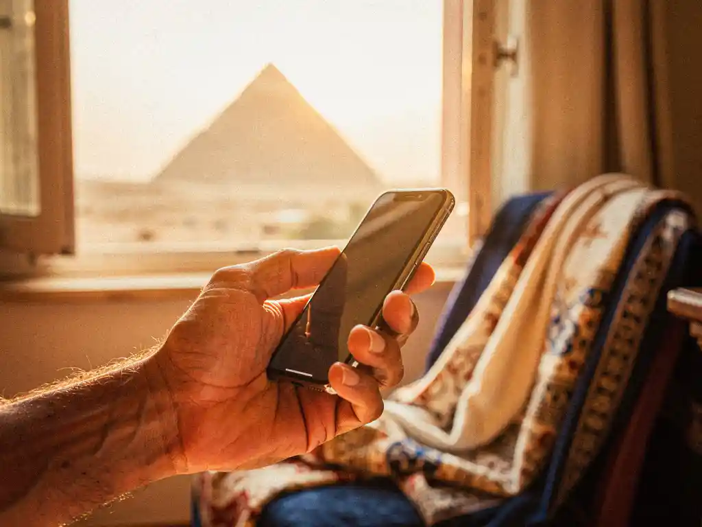 Weathered hand holding a smartphone near a sunlit window with a faint pyramid silhouette, Arabic textile draped alongside in warm terracotta tones.