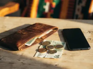 Worn leather wallet open on a sunlit wooden table with euro coins, a folded banknote, and a smartphone beside kente fabric.