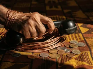 Weathered hand gripping a vintage rotary phone receiver tangled in copper wire coiled around multinational coins on a kente-cloth surface.