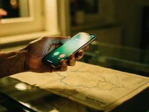 Weathered hand holding a smartphone with a glowing call screen, faded South Sudan map beneath glass desk, warm amber dusk light through apartment window.