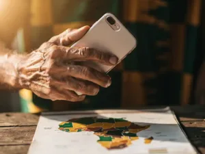 Weathered hand holding a smartphone to the ear, warm Kente-inspired amber and green bokeh, blurred West Africa map on wooden surface below.